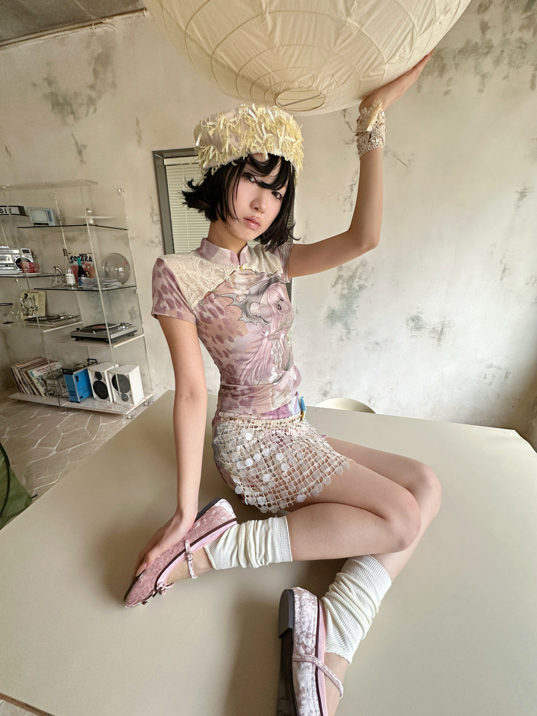 4RE1GN DREAM GARDEN sequin-panel mini skirt in pink multicolor — seated modeled view with one hand raised to a paper lantern