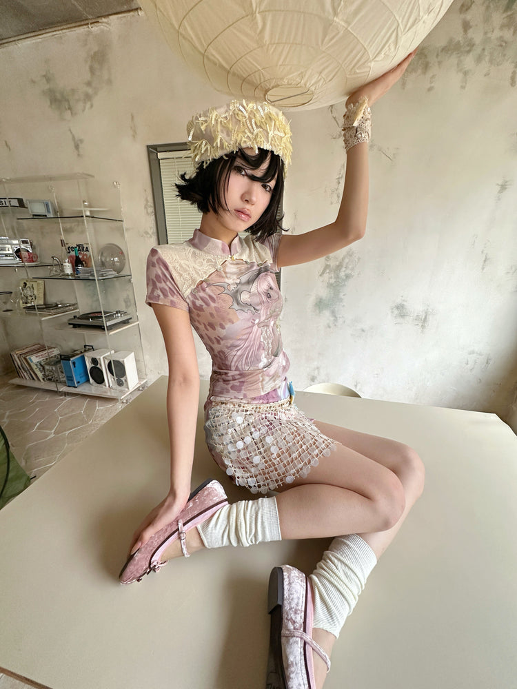 4RE1GN DREAM GARDEN sequin-panel mini skirt in pink multicolor — seated modeled view with one hand raised to a paper lantern