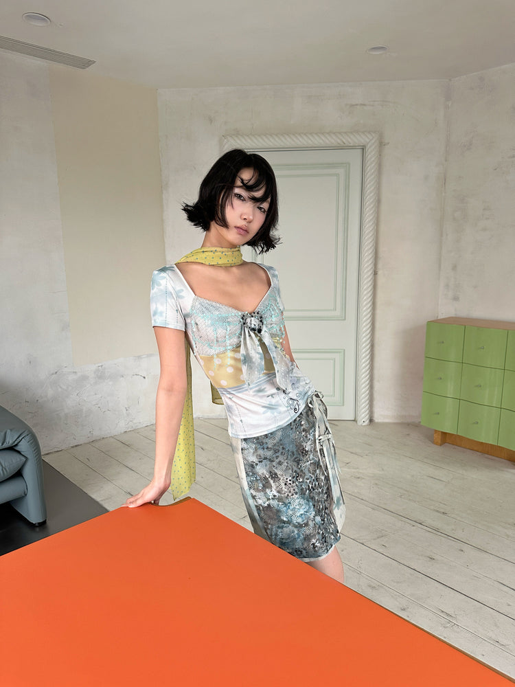 4RE1GN BLUE BLOOM patchwork short-sleeve tee in blue multicolor — modeled view seated on an orange table with a floral skirt