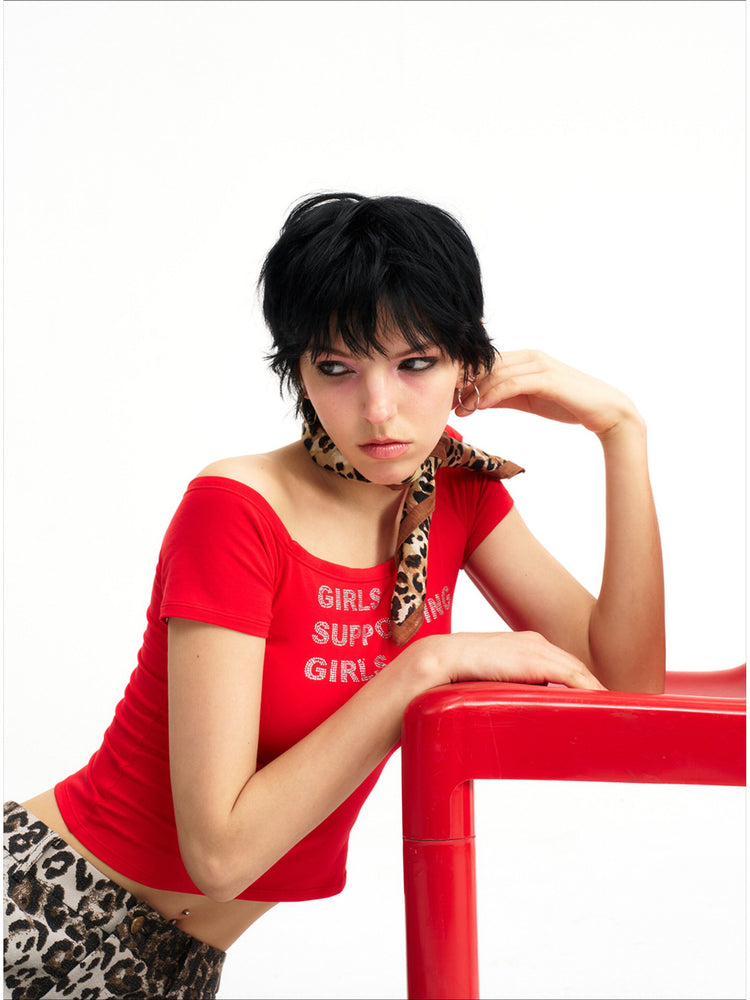 NEXT PASSPORT GIRLS SUPPORTING GIRLS rhinestone off-shoulder crop tee — red three-quarter modeled view beside a red chair.