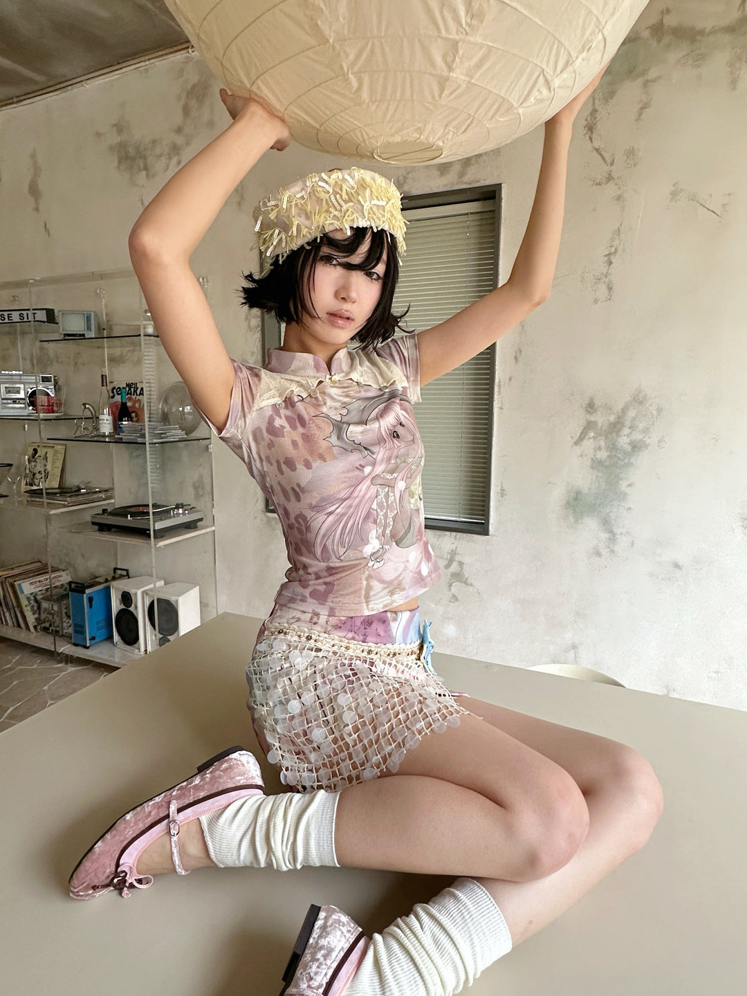4RE1GN DREAM GARDEN sequin-panel mini skirt in pink multicolor — seated modeled view with both arms raised to a paper lantern