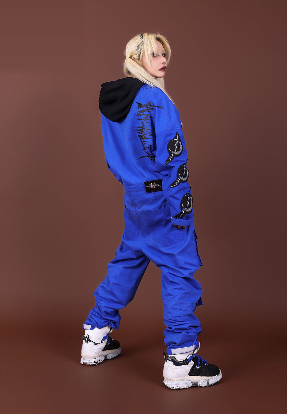 Rear three-quarter view of the blue REMAKE jumpsuit, with sleeve appliqués, rear pockets and a black hoodie styled underneath