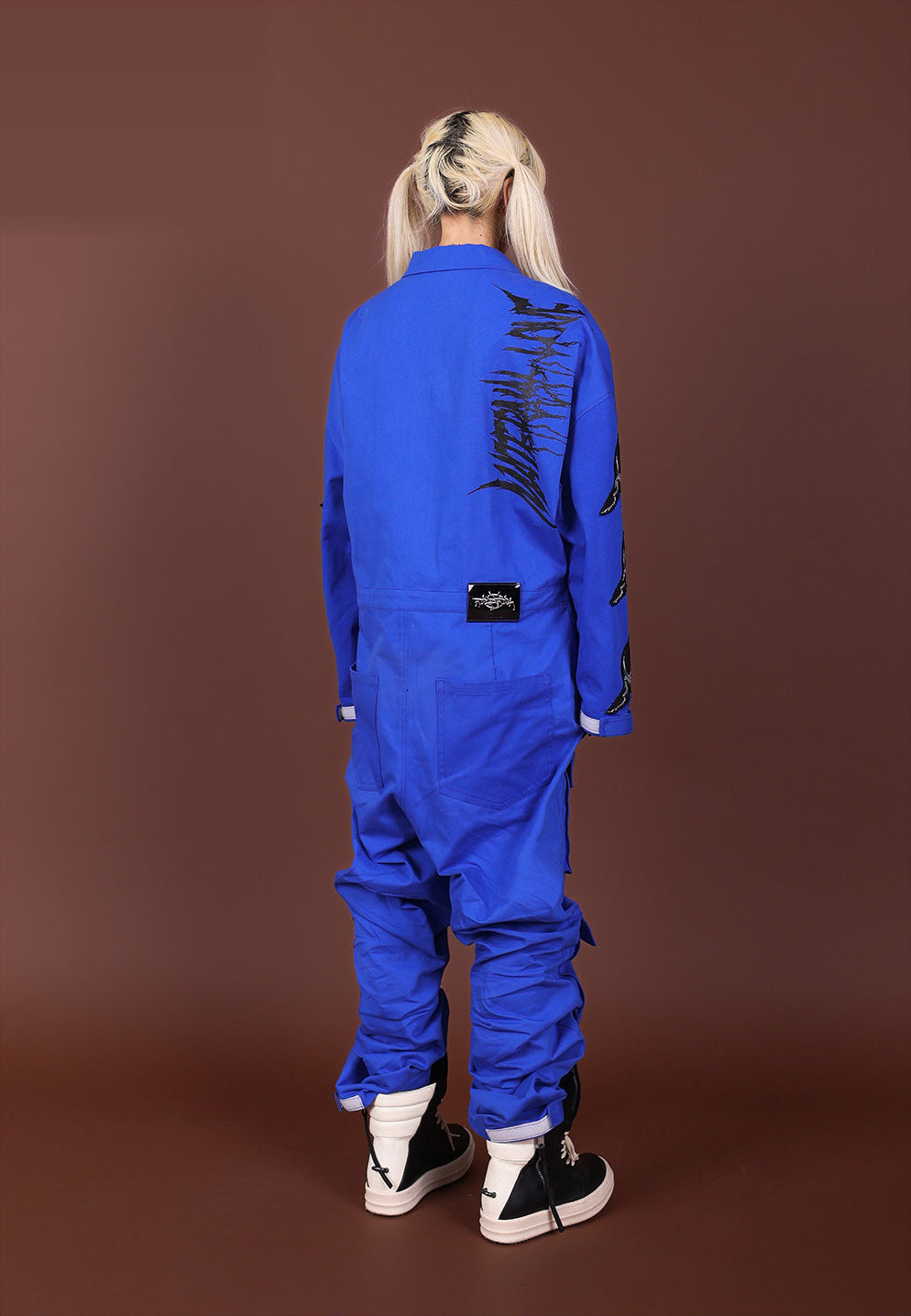 Back view of the blue REMAKE utility jumpsuit with black shoulder graphic, waist label and rear pockets