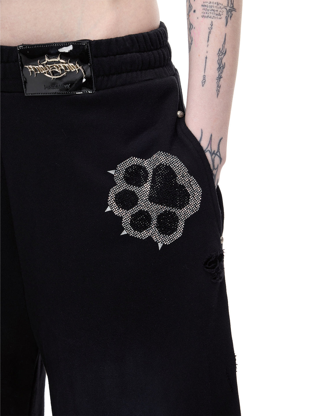 PINKSPINK CATPAW rhinestone curved-leg sweatpants in black — close front detail of the elastic waistband, metal logo patch and rhinestone paw