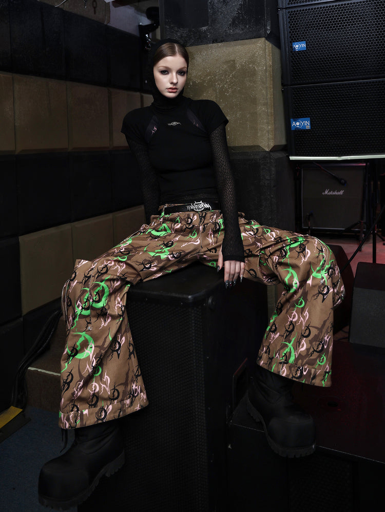 PINKSPINK ELECTRIC MOON sculpted camo cargo trousers in CAMOUFLAGE — seated front showing wide legs and flared hems.