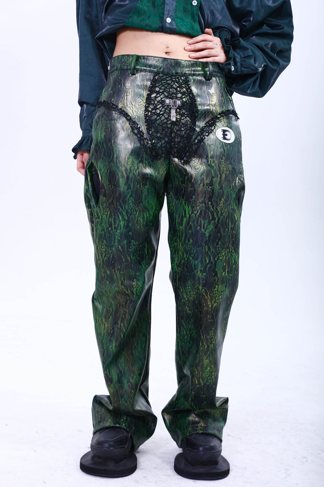 EMBRYO TRIANGLE ARMOR lace-inset leather pants in green — front modeled view with the overshirt lifted to show the waist construction