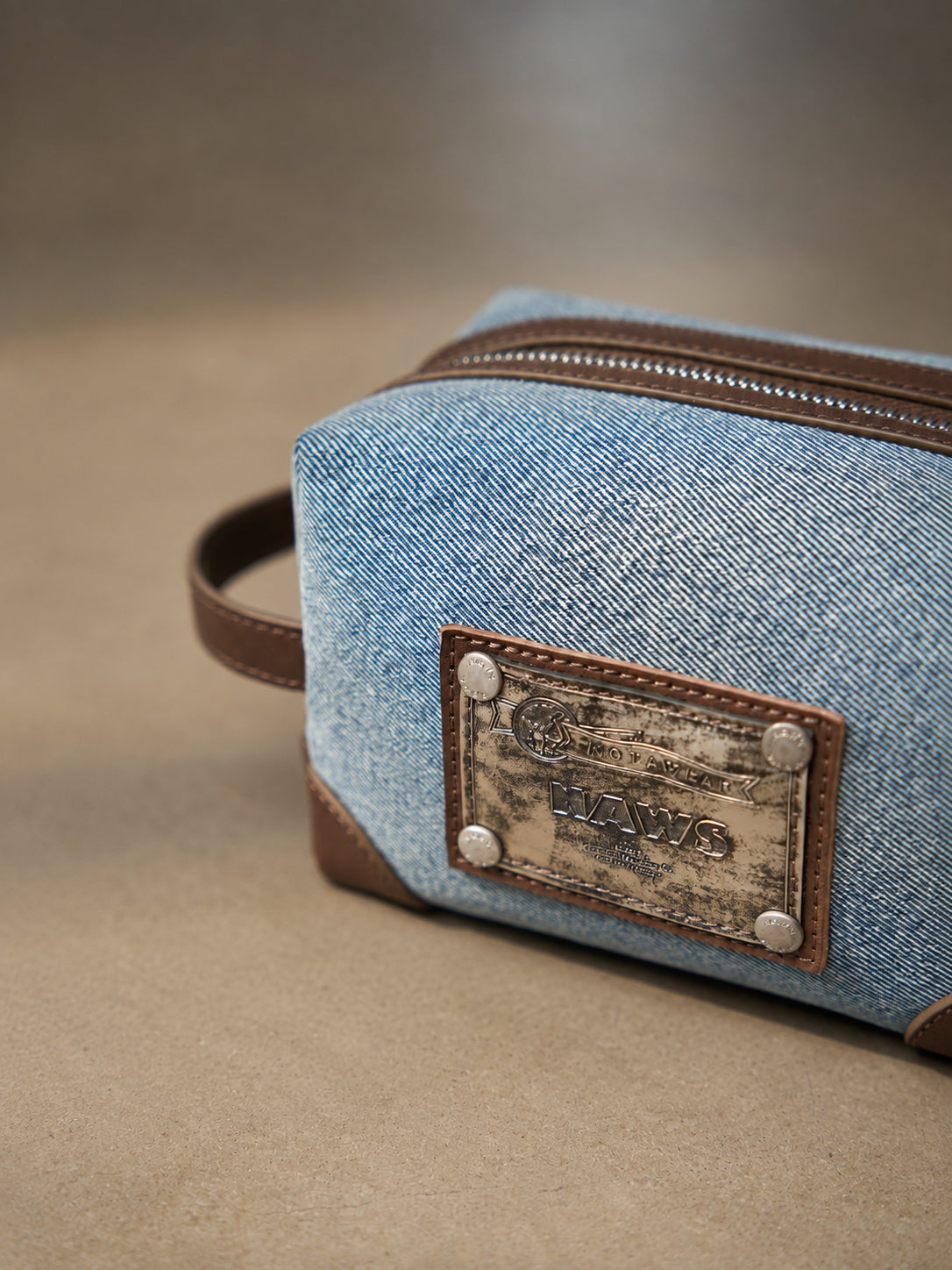 NAWS BLUE BOX denim makeup bag — close-up of washed denim, brown trim and weathered metal logo plaque