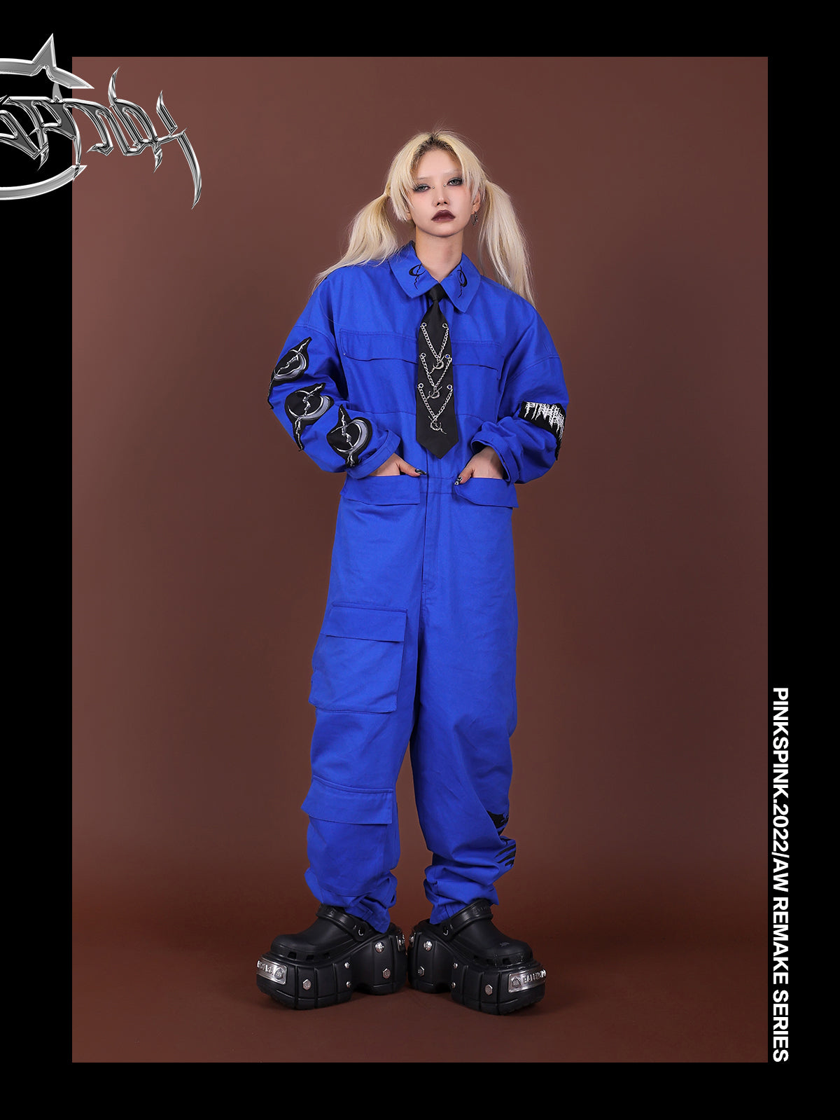 Blue REMAKE jumpsuit styled with a black chain-detail tie and both hands in waist pockets