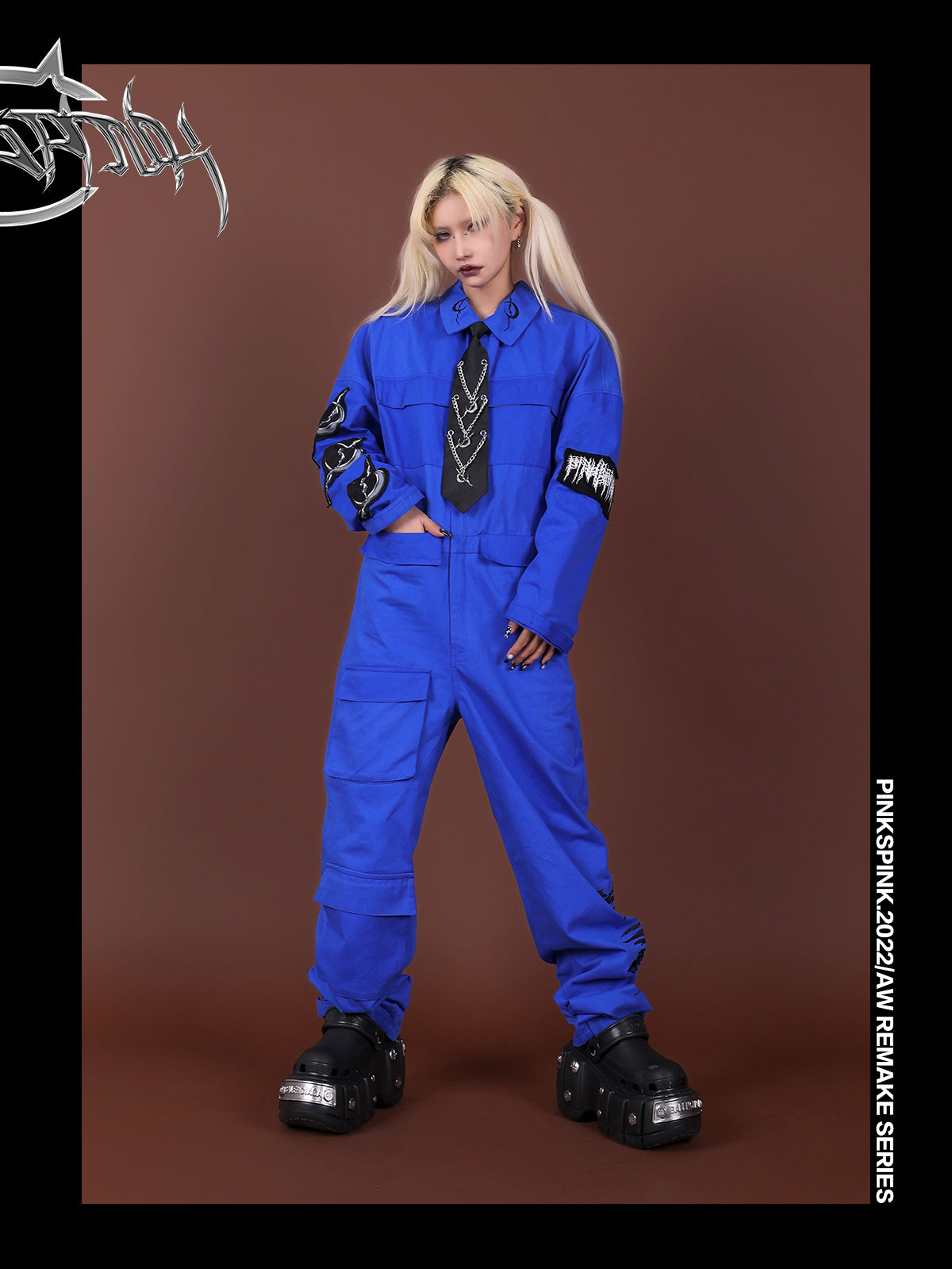 Blue REMAKE jumpsuit styled with a black chain-detail tie, one hand in the waist pocket