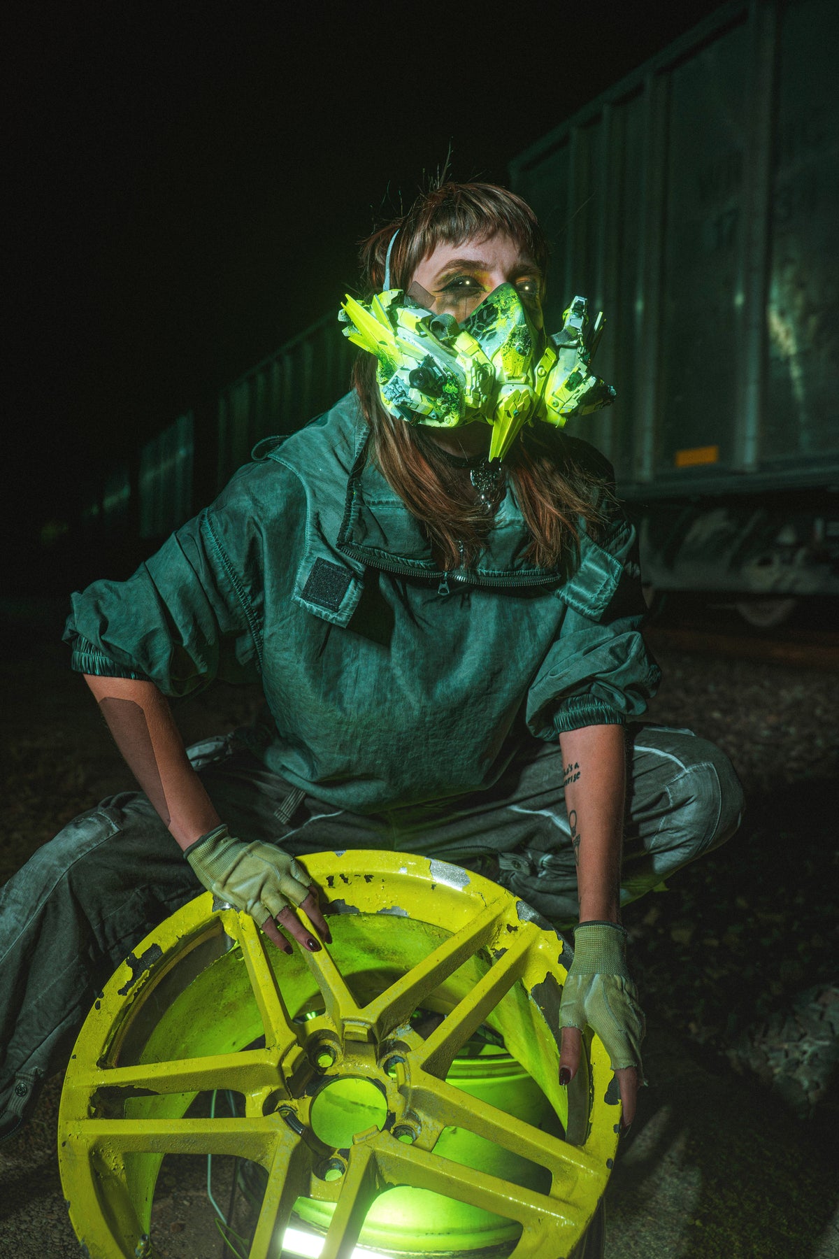 Julia in tactical futurewear beside a rail wheel at night
