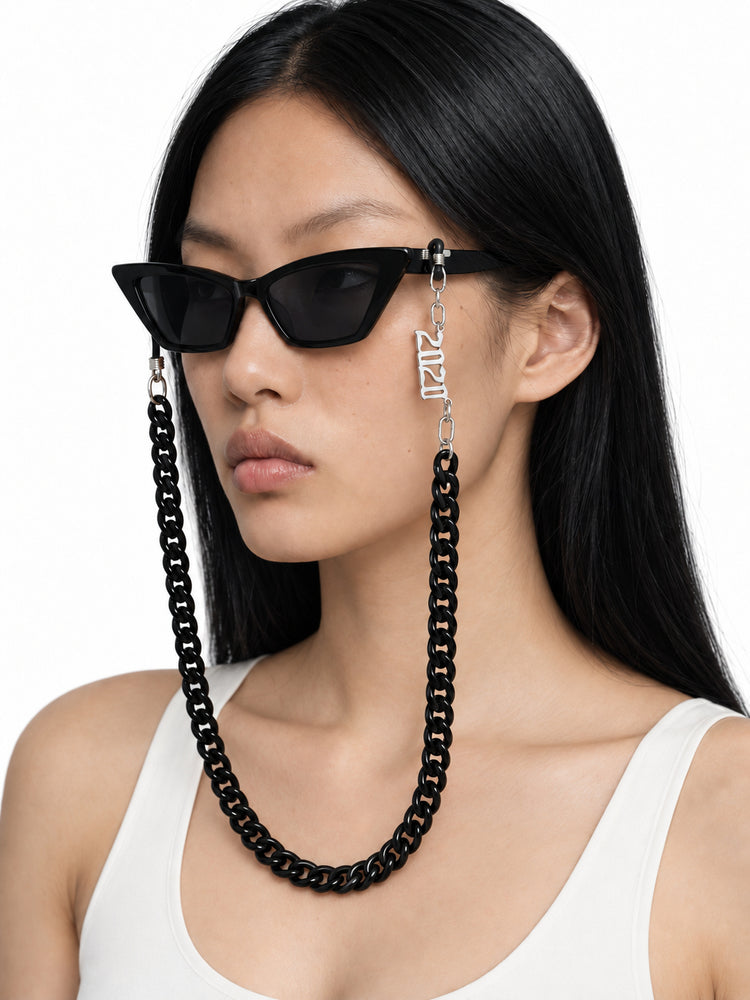 RICH BABY black cat-eye sunglasses with the included black chain, worn by Chell in a three-quarter studio portrait